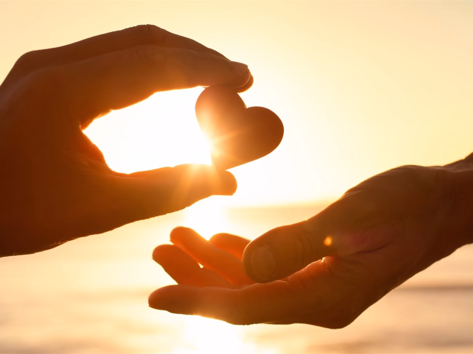 One hand passing a heart to another at sunset